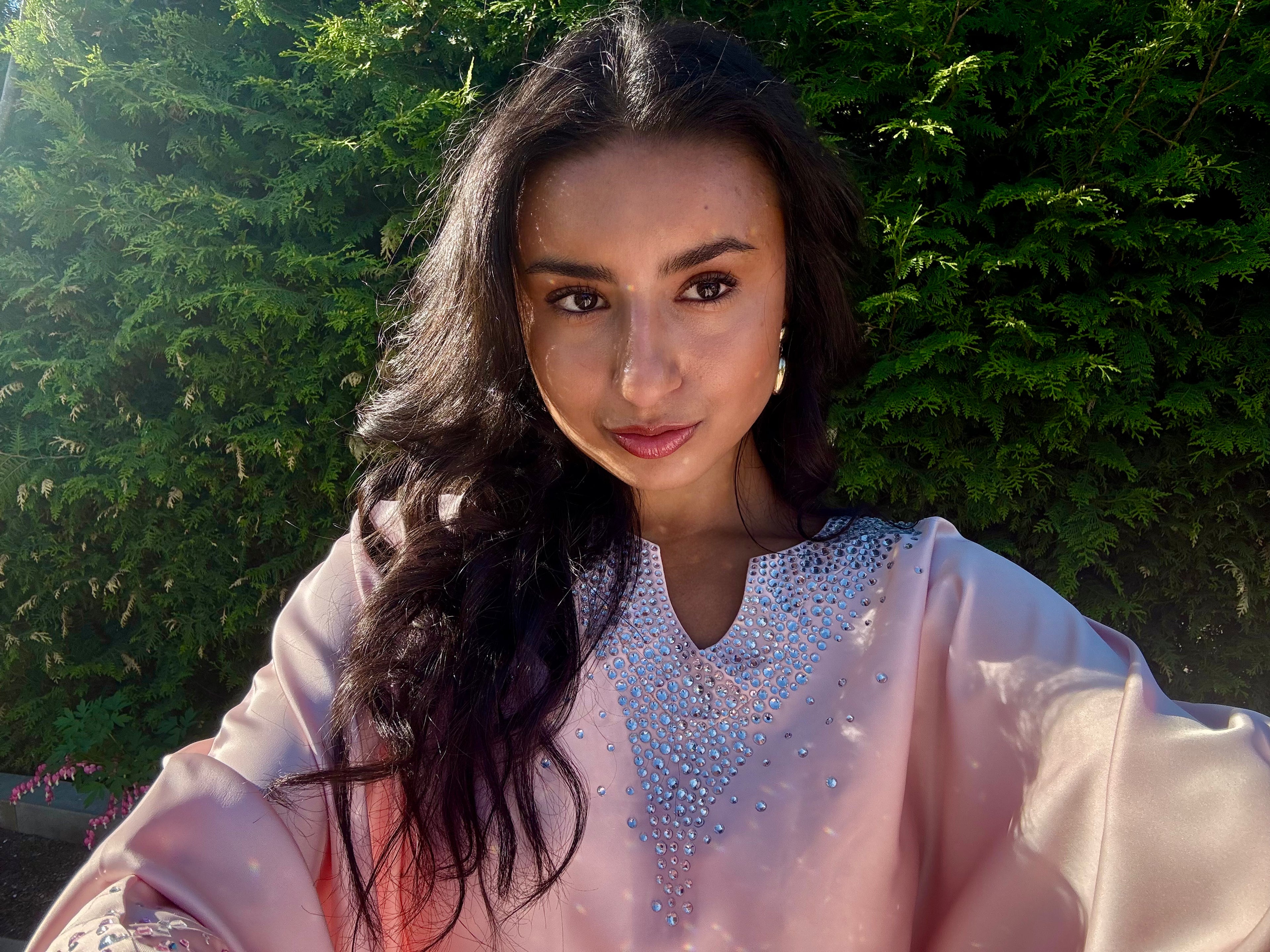 A woman with long dark hair wearing a light pink traditional-style top with a sparkling silver embellished neckline, photographed outdoors against a background of green foliage.